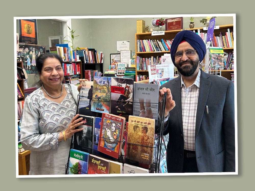 Amrit Kaur Chilana, left, is a Punjabi woman in her 60s with dark hair pulled back and a silver suit with a scalloped textured pattern. She stands on the left side of a black wire-framed bookshelf across from Rajwant Singh Chilana, right, a Punjabi man in his 60s with a navy turban, glasses and a dark blazer over a white shirt with a burgundy grid pattern. They are standing inside a crowded bookstore with many books on the shelves.
