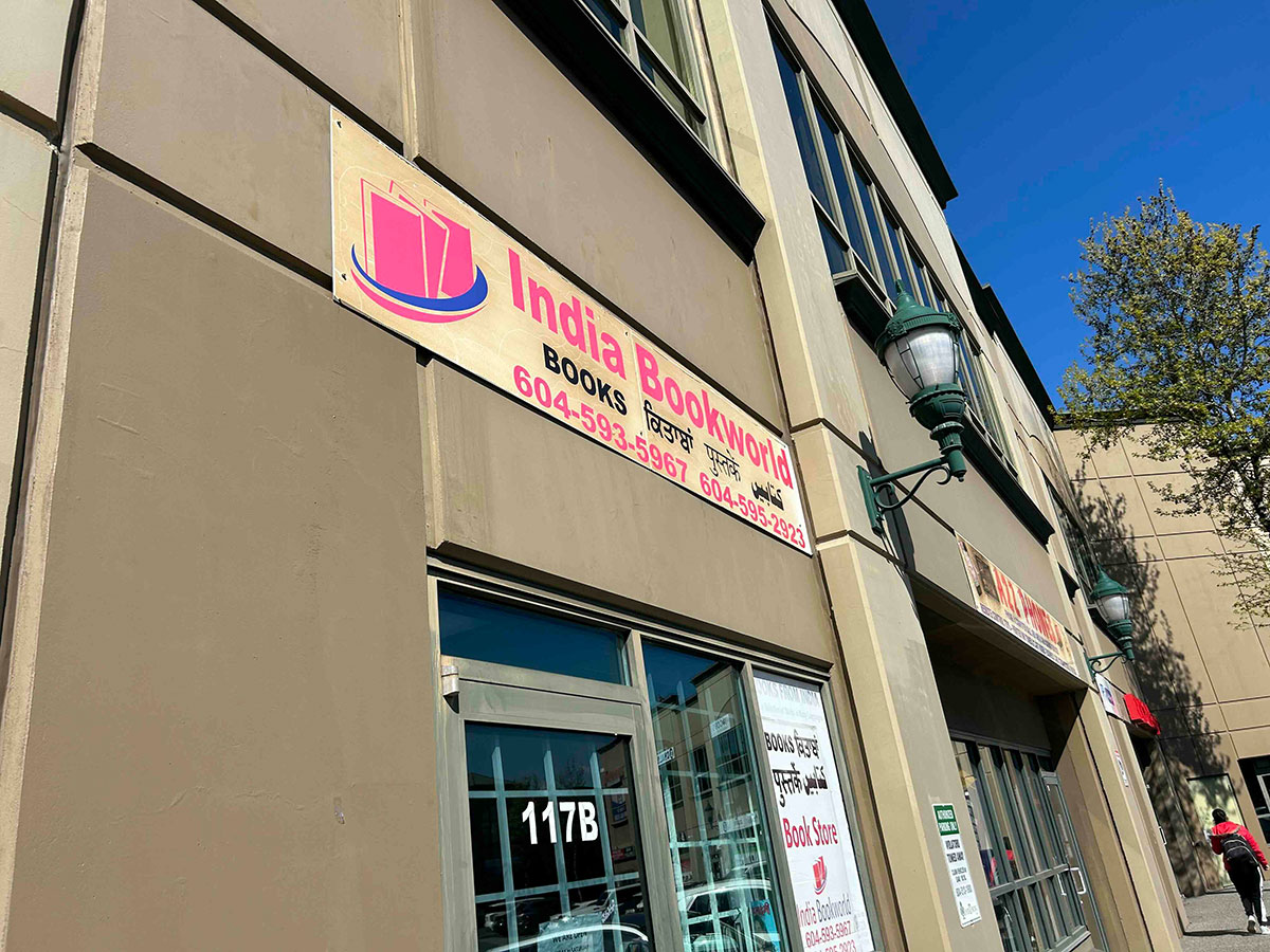 A beige storefront features a banner with neon letter sans-serif text that reads 'India Bookworld' over a modest doorway on a sunny day. The shop is part of a retail complex, with adjacent shops part of the same building.