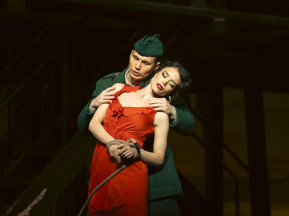 Two opera singers stand onstage against a darkened backdrop. Matthew White, left, has short hair, a green beret and light skin. He is clad in a green military costume. He holds the shoulders of Ginger Costa-Jackson, right, who leans against him with her eyes closed. She has light skin, dark wavy hair and dramatic makeup. She wears a red dress and her wrists are tied with ropes.