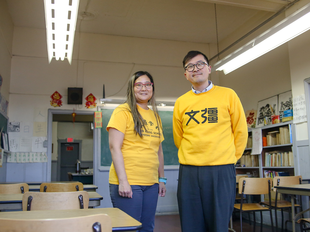 Aynsley Wong, left, and Jeffrey Wong, right, are clad in yellow Wongs’ Benevolent Association T-shirts. They are both younger Chinese adults with light skin and glasses. They’re standing in a classroom with rows of birch desks. A green chalkboard is in the background.