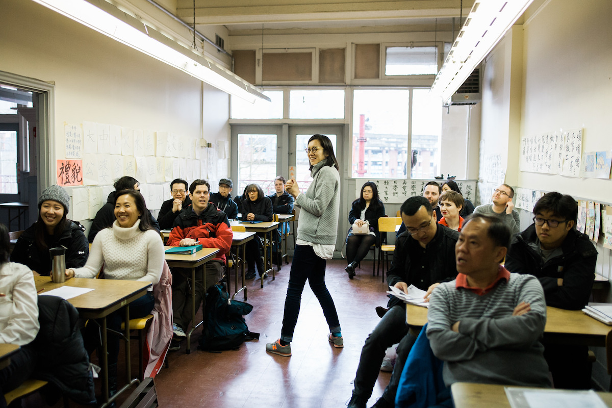 A classroom features two aisles of two-person desks filled with adults. June Chow stands between the aisles in a grey sweater and black pants. She is a Chinese woman with shoulder-length black hair and glasses.
