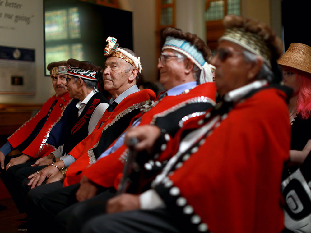 Five Indigenous men in regalia, including red and blue cloaks and head wear, sit in a row.