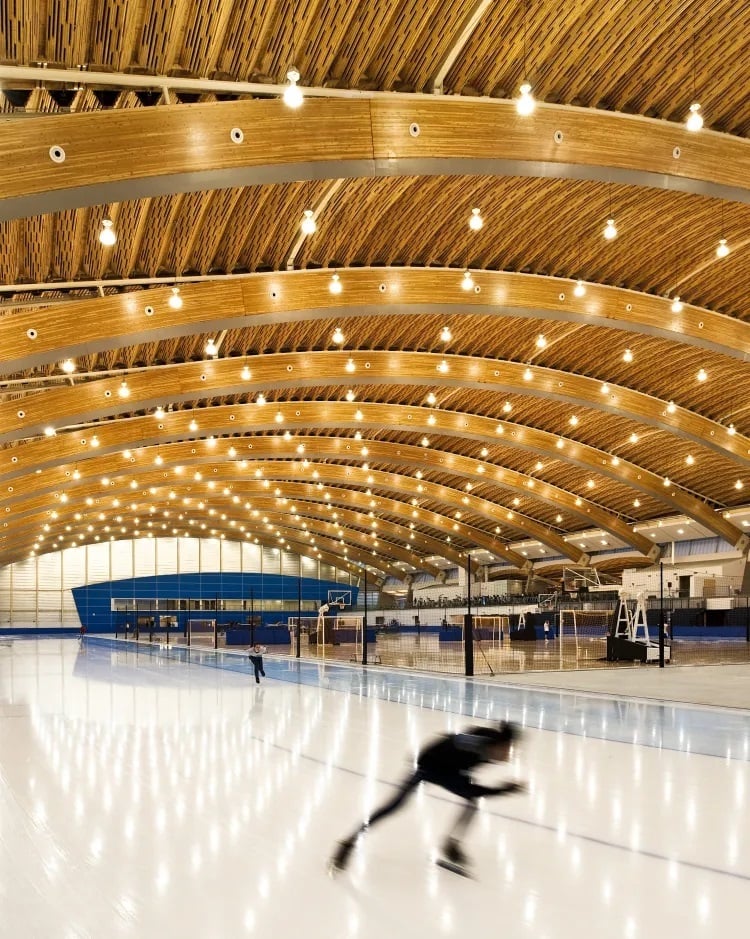A skater on ice in an arena under an arching wooden roof with many components.