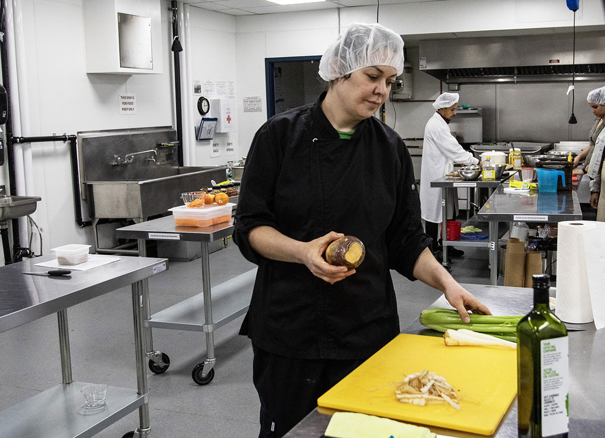 A woman with medium-light skin wearing a white hair net and a black chef's jacket stands in a kitchen, holding a rutabaga in one hand. On the stainless steel counter in front of her is a bunch of celery and a couple of peeled parsnips on a yellow cutting board.