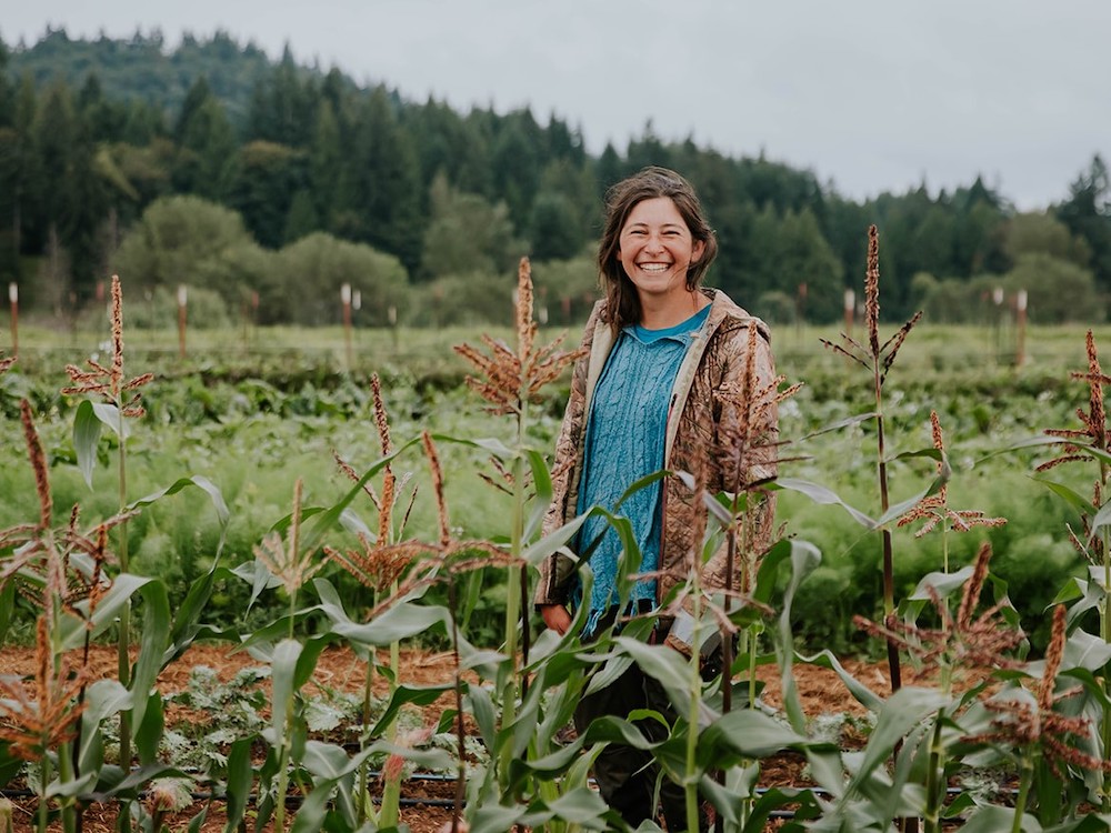 A smiling woman stands in a productive-looking farm field. She has shoulder-length dark hair and wears a light blue sweater and open jacket.