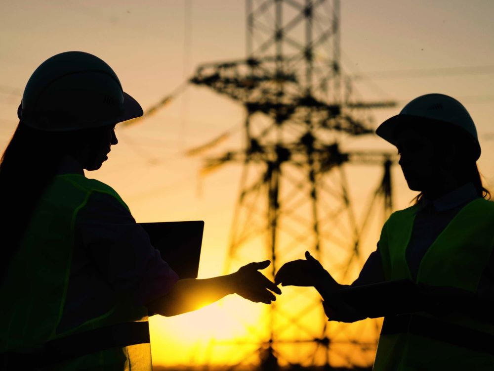 Two power engineers in hard hats and vests gesture to shake hands against a backdrop of an electrical transmission line at sunset. They have long hair and the person on the left is holding what looks like a clipboard. The sky is orange and blue.