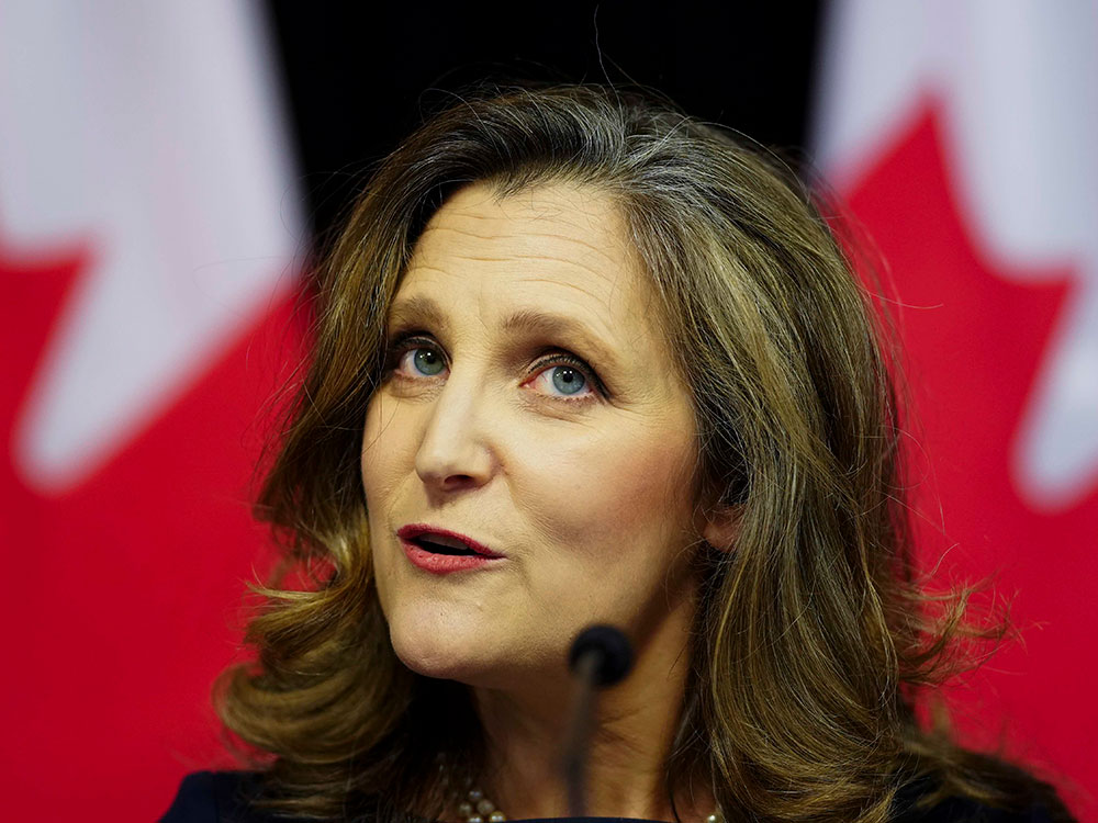 A close-up of a 55-year-old woman with brown hair streaked with grey stands. She wears red lipstick and is in front of an out-of-focus Canadian flag.