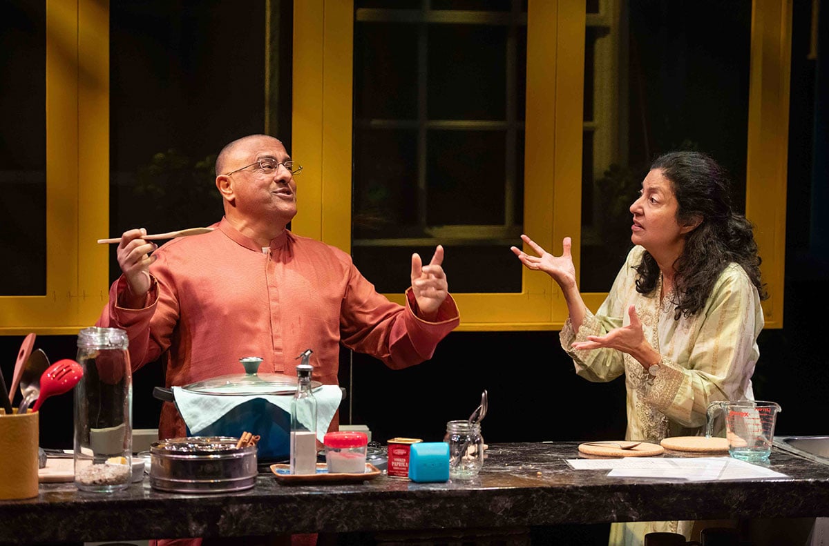 Two middle-aged actors stand behind a kitchen counter onstage under bright lights. On the left, Parm Soor raises his hands and a wooden spoon mid-speech. He is bald, has glasses and wears a light orange button-down shirt. On the right, Nimet Kanji gestures towards him. She has long wavy dark hair and is wearing a light yellow dress.