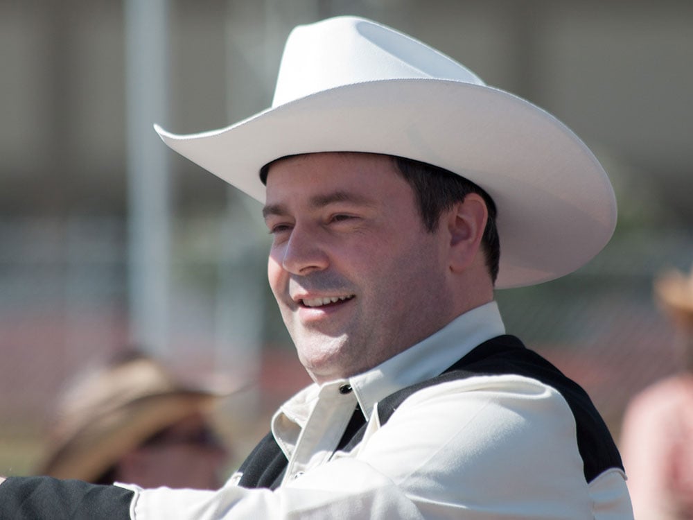 A white man in his 40s smiles while wearing a bright white cowboy hat and black and white cowboy-styled shirt.