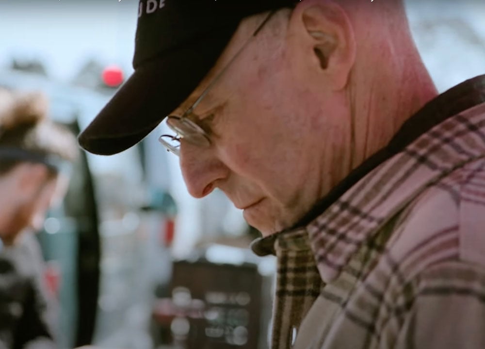 A middle-aged man in glasses, a dark ball cap and a plaid jacket looks down to the left of the frame. In the background in soft focus are people at a farmers market.