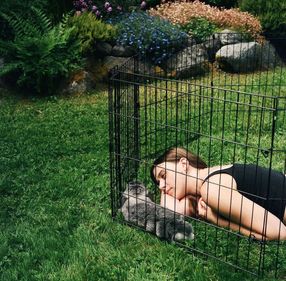 A young woman with light skin and brown hair lies on her stomach in the green grass inside a black cage for pets. She is wearing a black tank top. Next to her is a small grey rabbit.