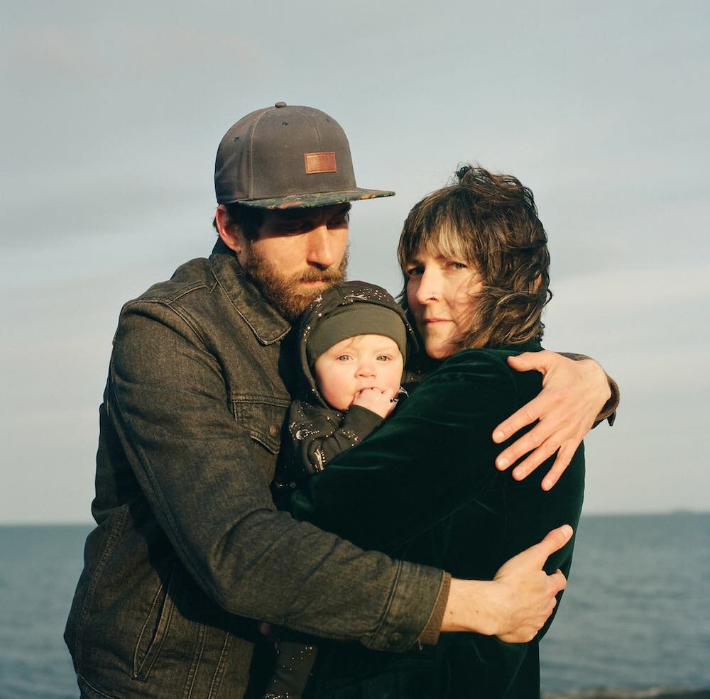 A woman with light skin and short brown hair looks towards the camera. She is holding a baby. A man with light skin, a ball cap and beard wraps his arms around them. In the background is the ocean reflecting a grey-blue sky.