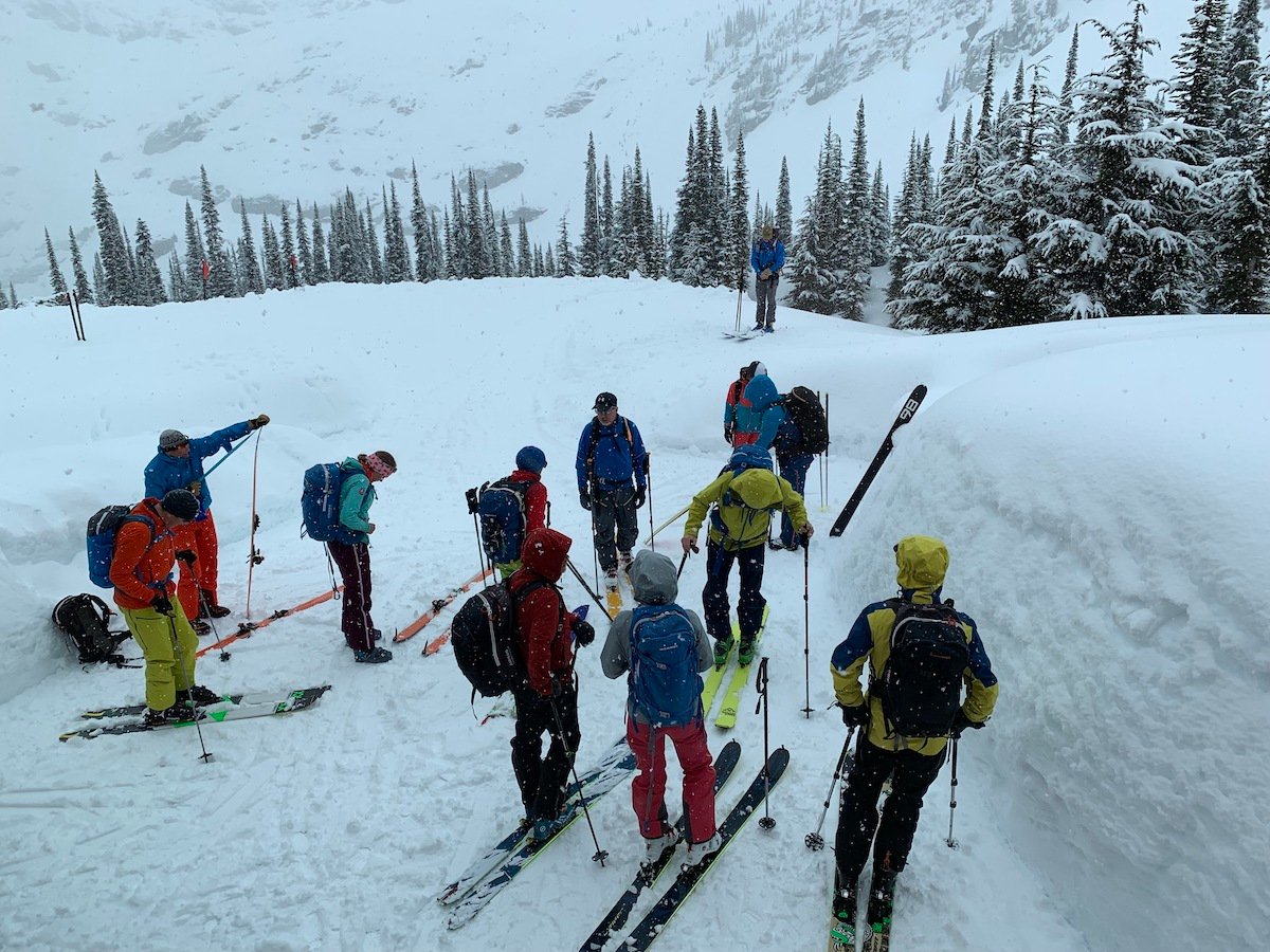 About a dozen people wearing skis and colourful winter clothes gather on a snowy slope. It is snowing.