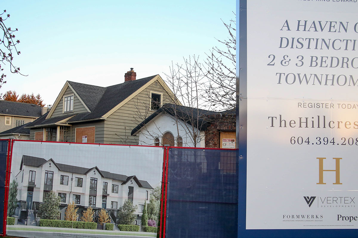 A close-up shot of a real estate advertisement featuring colour photos of white townhomes wraps around a construction fence. Behind it are boarded-up single-family homes, one sage green and one white, against a cool blue sky.