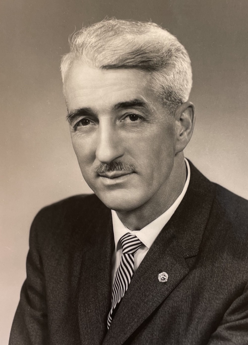 A black and white archive photograph of a light-skinned man with a slight smile wearing a dress shirt, a suit and a striped tie. He has white or grey hair and sports a moustache. He is wearing a pin on the left side of his chest.