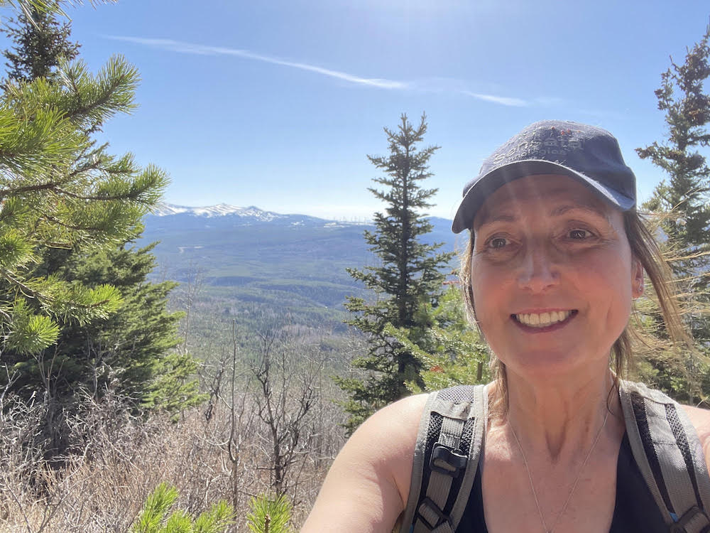 A woman pauses from hiking to take a selfie. Snowy mountains are in the background and she smiles at the camera.