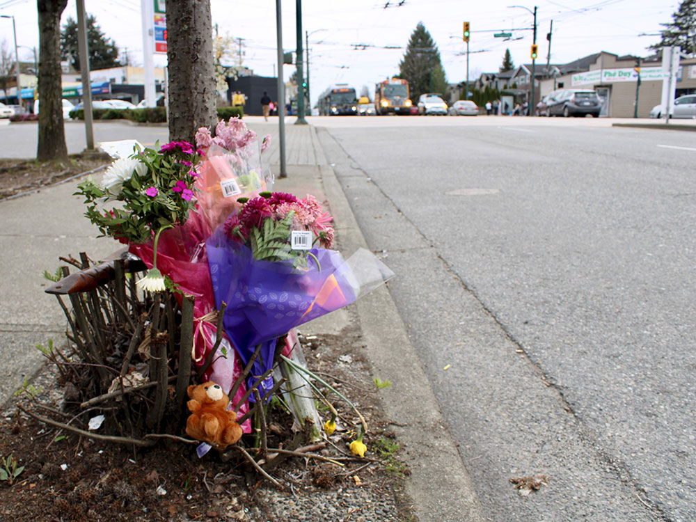 A few bouquets and a small teddy bear are placed beside a post on a sidewalk.