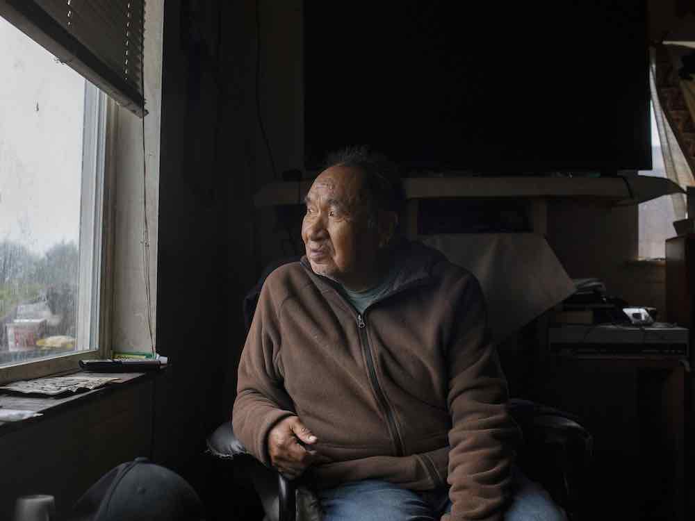 An elderly Indigenous man with medium skin tone looks to the left of the frame, out the window of his home. He is sitting in a dimly lit room wearing a brown zip-up hoodie.