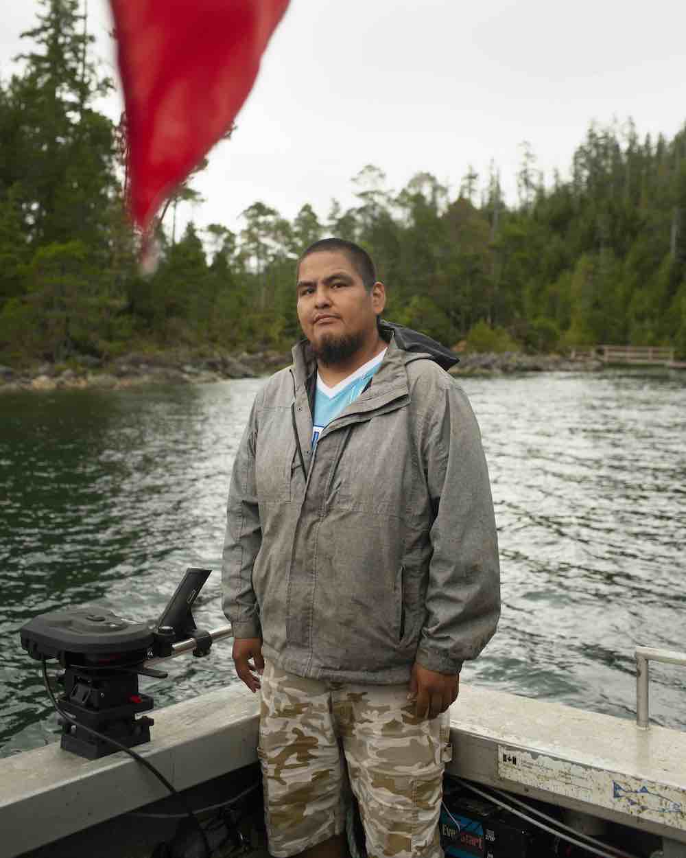 Sammy Williams stands aboard a fishing boat. He has medium skin tone, short dark hair in a buzz cut and a dark beard. He wears a grey rain jacket and beige camo shorts. A red flag hangs in the upper left quadrant of the foreground.