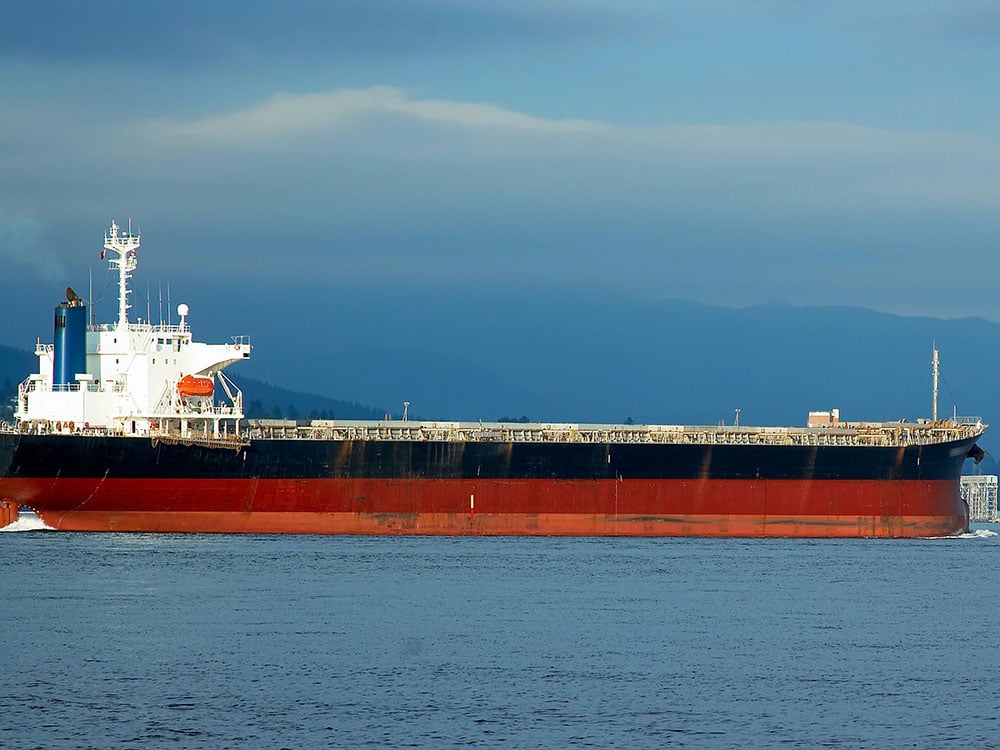 A large tanker with white superstructure and a white, red and orange hull sits in Burrard Inlet, with mountains in the background.