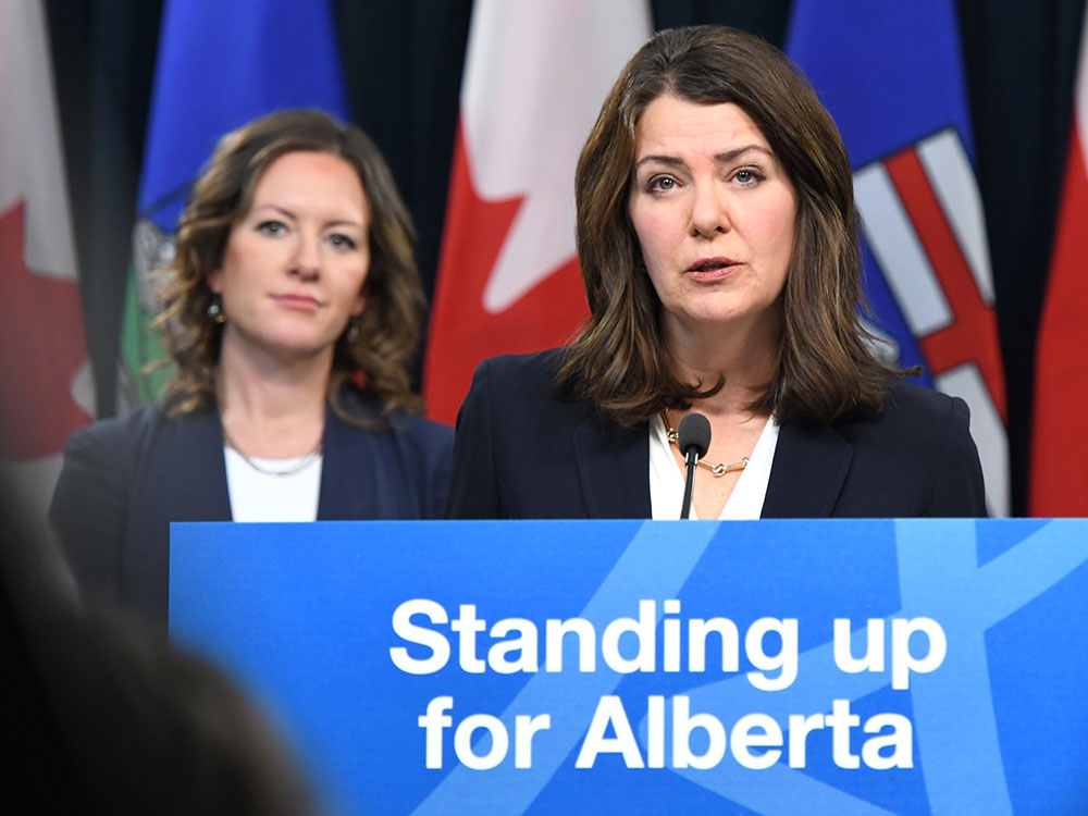 A light-skinned middle-aged woman stands at a podium with a blue sign that says 'Standing up for Alberta.' She has shoulder-length brown hair and wears a blue jacket and white shirt.