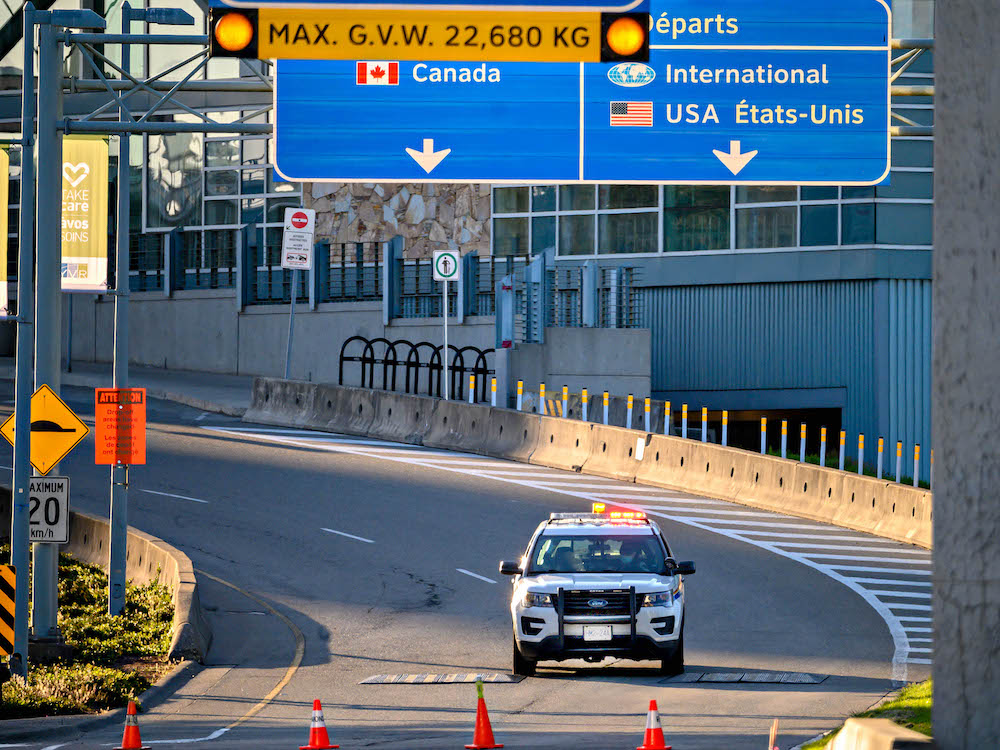 A white sports-utility vehicle with a thick black grill is parked with its front facing the camera before a row of orange pylons on a concrete ramp. Bright blue signs above the ramp say “Canada” on the left and “International” on the right, signalling to drivers which lane to use for accessing different airport departure terminals. 