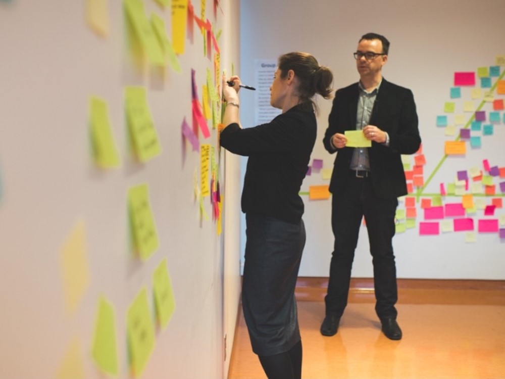 Julie Szabo, left, writes on a sticky note to go onto a wall full of sticky notes. Darren Barefoot, right, holds two more yellow sticky notes and looks on intently.