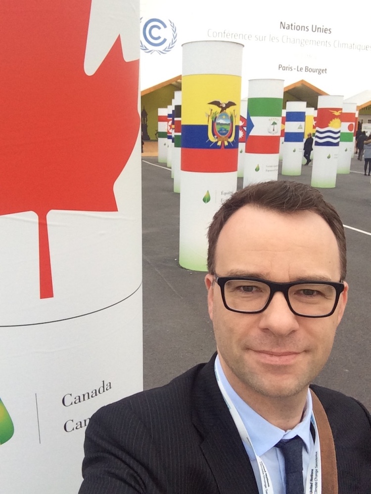 Darren Barefoot, who has light skin and short brown hair, stands in front of a series of pillars featuring different countries’ flags at COP21.