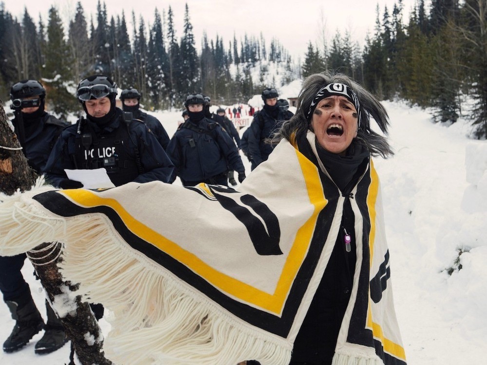 A woman with medium-light skin tone and dark grey hair, wearing a patterned headband and a fringed blanket wrapped around her shoulders, appears to be in mid-shout in a snowy, treed area. Behind her are several police officers in black uniforms, helmets and goggles.