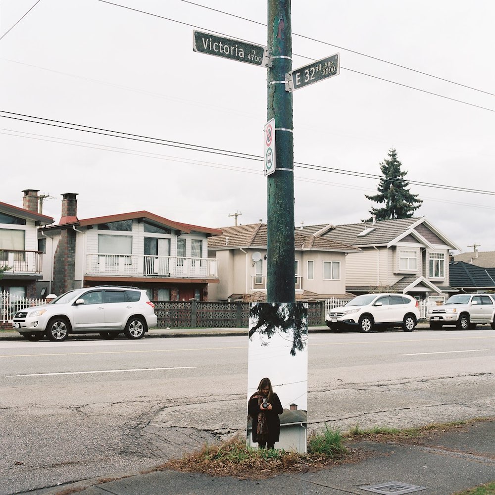 A green traffic pole bears black street signs with white text that read 'Victoria Drive' and 'East 32nd Avenue.' At the bottom of the pole leans a mirror against which the reflection of Jackie Dives can be seen; she is wearing a dark jacket, looking down and holding a camera against her chest. Behind the pole are rows of Vancouver Special and other houses on a residential block with parked cars on the side of the road. The sky is grey.