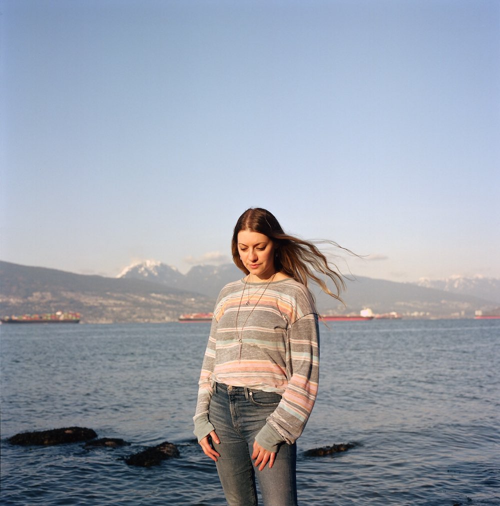 A young woman with light skin and long, wavy, light brown hair stands on a beach in a pink and grey striped sweater and jeans, looking down. Behind her is the ocean, ships and Vancouver’s mountains in the background. The sky and water are blue.