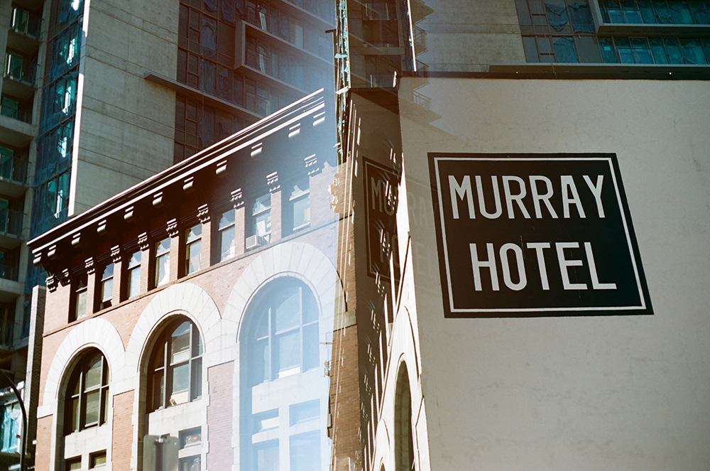 A double-exposure image of a grey building on a bright day with a black sign and white text that reads 'Murray Hotel.'