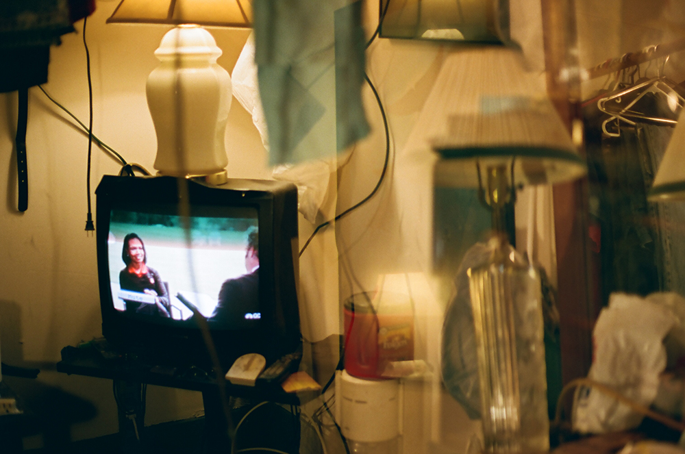 A double-exposure image depicts, on the left, a small black television with a white lamp perched atop it. Onscreen, a woman in a black suit is being interviewed by a man in a black suit. On the right are coffee containers, a transparent glass lamp and clothes hangers.