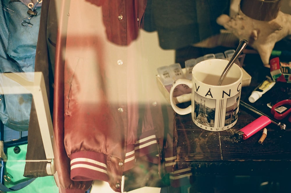 A double-exposure image depicts, on the left, a row of jackets including a burgundy baseball jacket, and, on the right, a white coffee mug with the word 'Vancouver' and black and white photos of the city on it. The mug sits on a table of assorted items including a pillbox, a cigarette and a pink lighter.