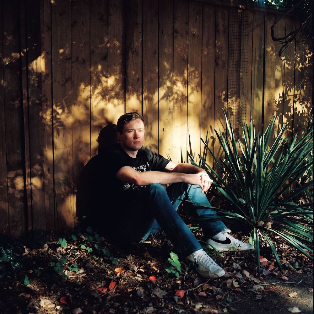 A young man with light skin and short blond hair sits on the ground against a wooden fence, refracted sun on his face. He is wearing a black T-shirt and blue jeans with white Nike sneakers.