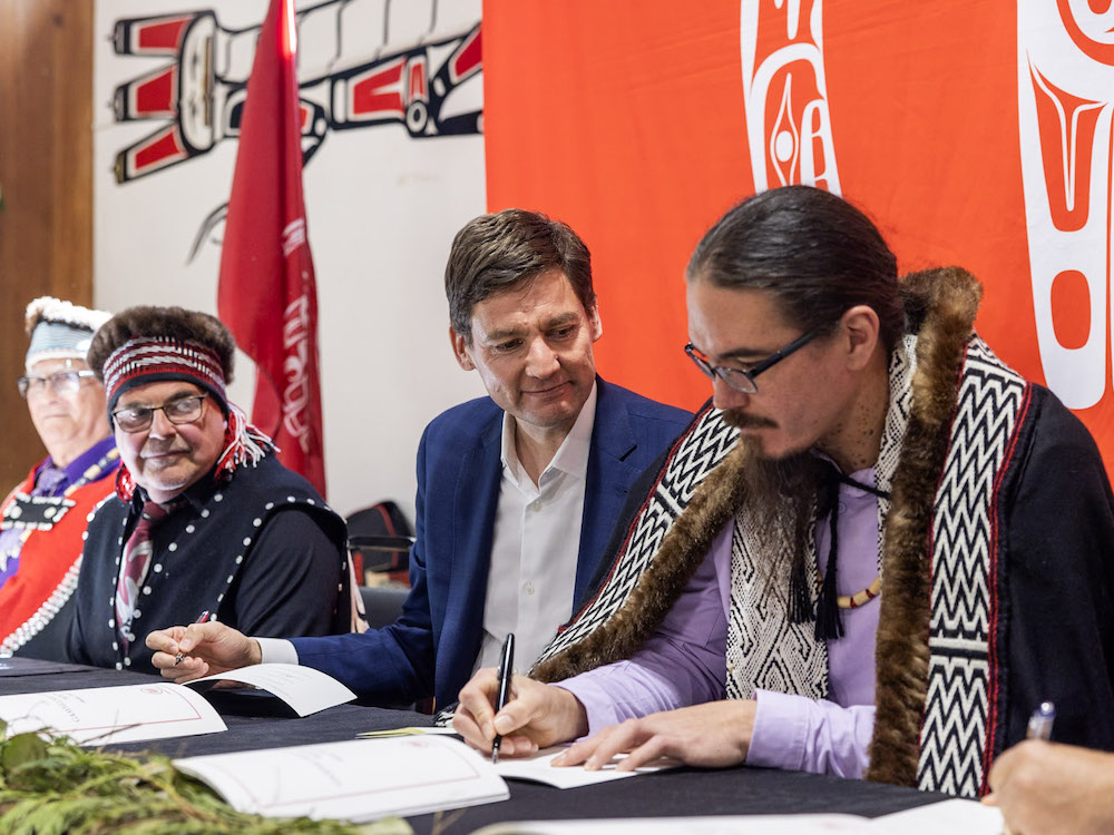 Four men sit at a table, signing documents. The members of the Haida Nation are wearing elements of traditional Haida dress. Premier David Eby is wearing a blue blazer and a white shirt.