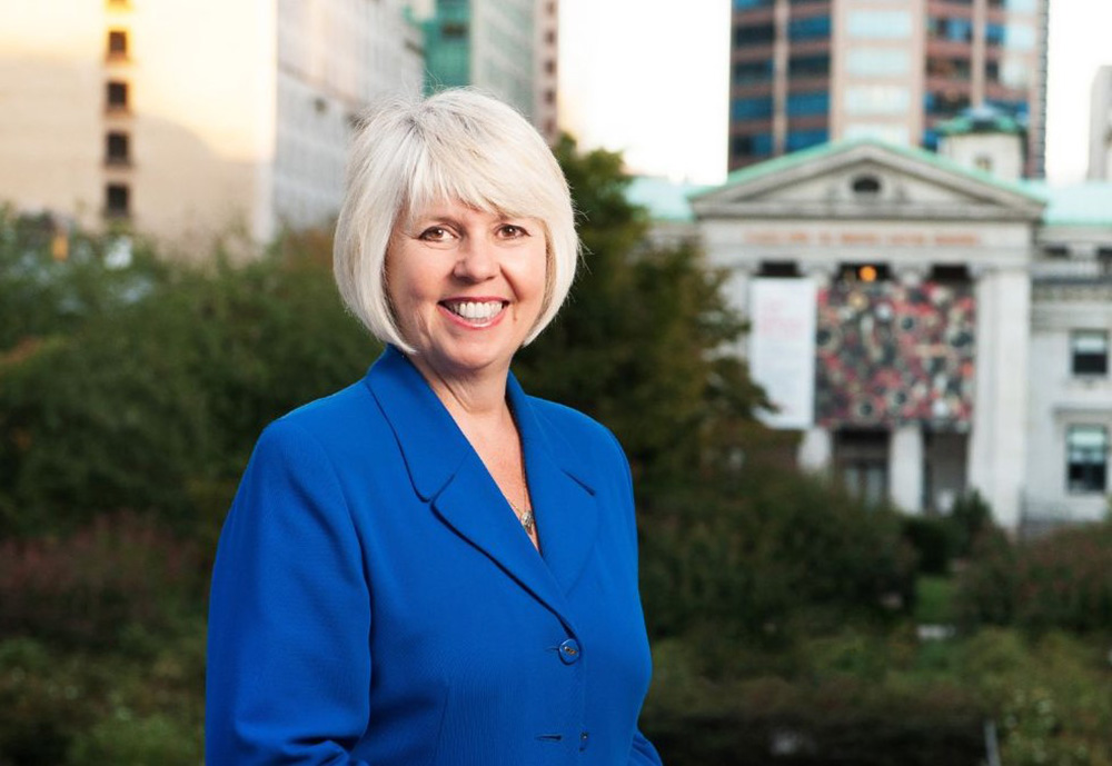 Adriane Carr has short white hair in a bob and bangs. She is smiling at the camera and is wearing a royal-blue blazer. She is standing outdoors against green shrubbery. In the background is the Vancouver Art Gallery.