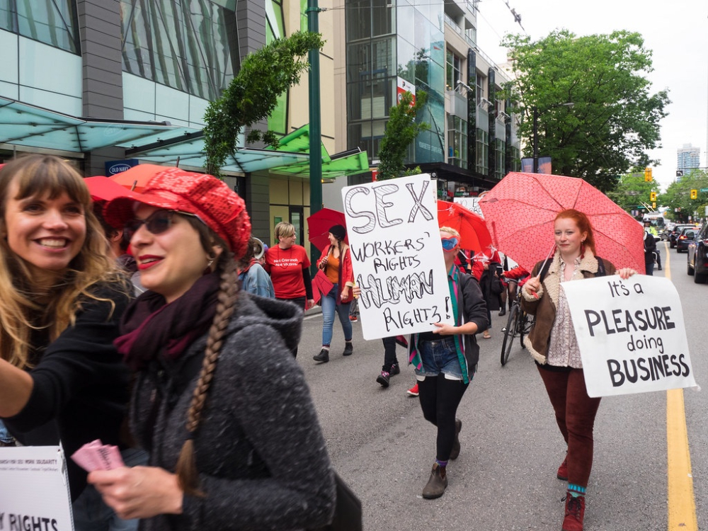 Women wearing red hats and carrying red umbrellas walk in a street protest. A sign reads &ldquo;Sex Workers&rsquo; Rights, Human Rights.&rdquo;
