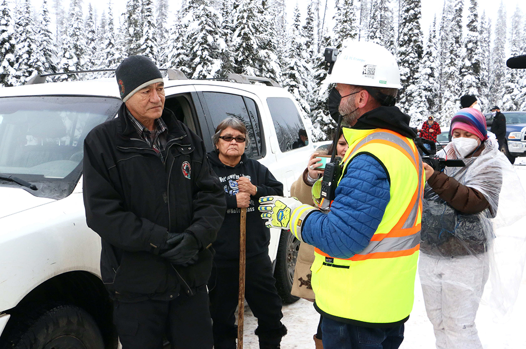 A man with medium skin tone, wearing a black toque, a black jacket and black gloves, stands outside a white SUV listening to a worker with light skin tone wearing a bright yellow safety vest, a white hard hat and a black medical mask. It is snowy out.
