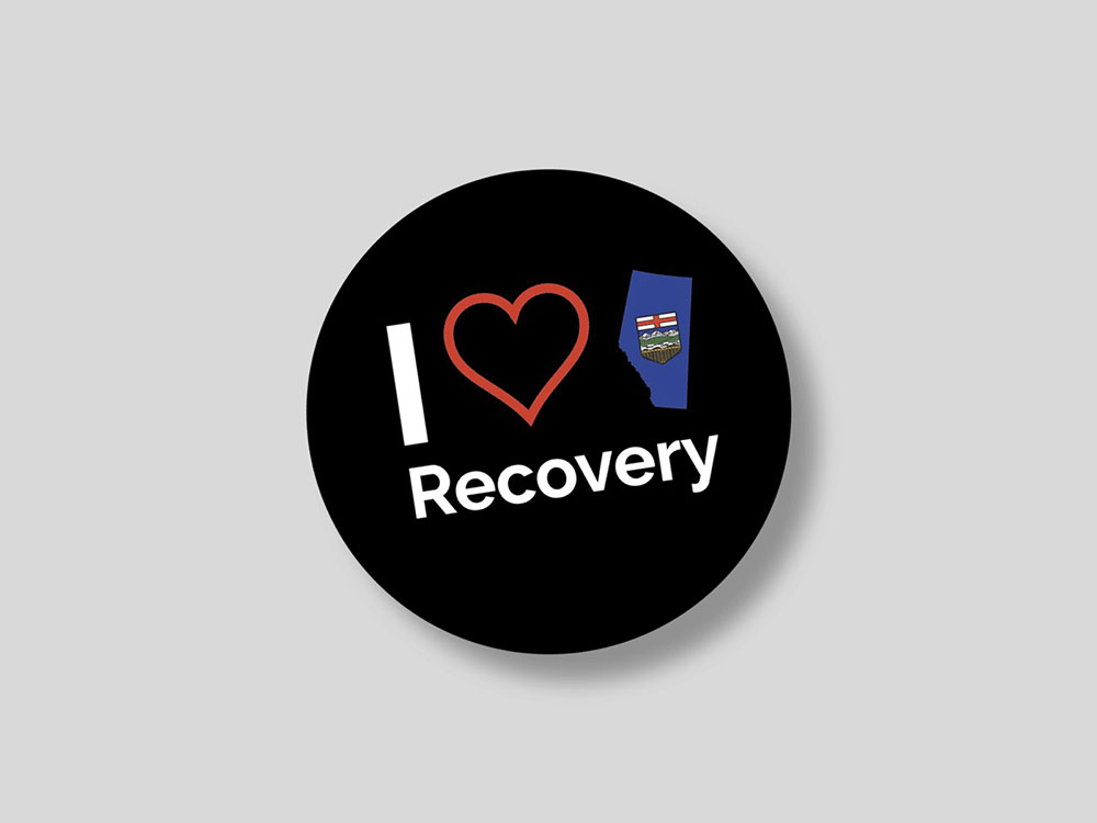 A black circular shape on a grey background reads &ldquo;I heart recovery&rdquo; in white text. The heart is an illustrated red outline of a heart next to a blue cut-out of the province of Alberta as it would appear on a map, with its provincial crest at the centre.