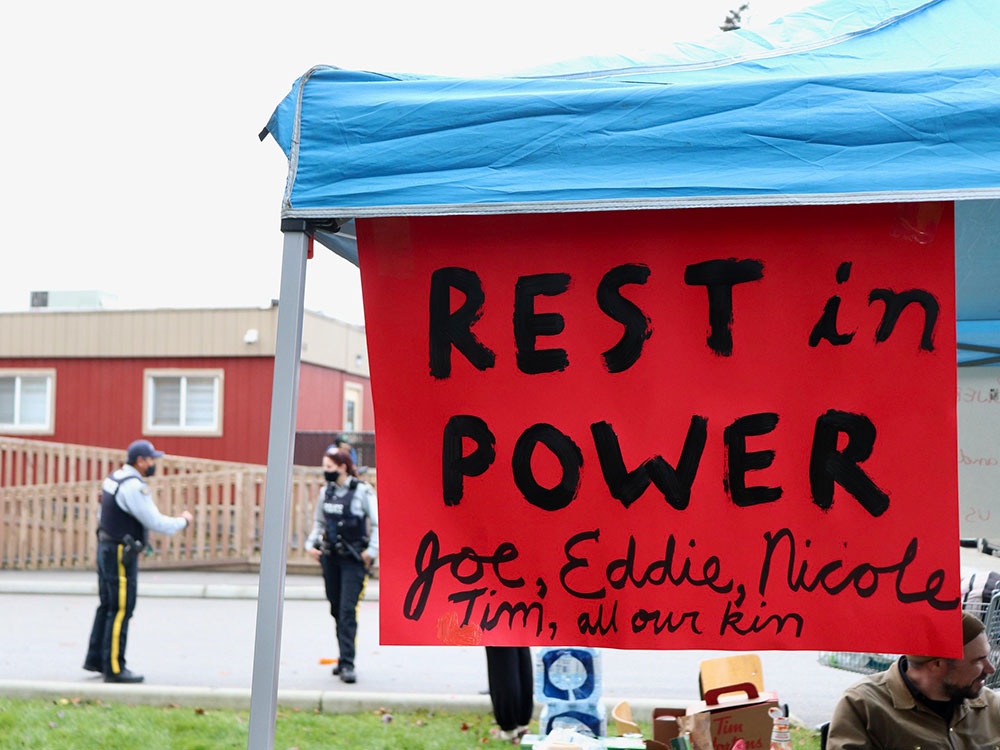 A handmade red sign hangs from the roof of a folding outdoor tent with a blue tarp. Hand-painted letters read, &ldquo;Rest in Power Joe, Eddie, Nicole, Tim, all our kin.&rdquo; A person in a toque and tan jacket is sitting to the right of the frame under the tent. Two police officers in medical masks are speaking with each other on the street in the background.