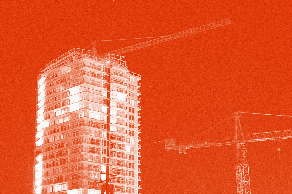 A condo building with some lighted up windows and some dark windows stands against a red background. Two construction cranes can be seen in the background.