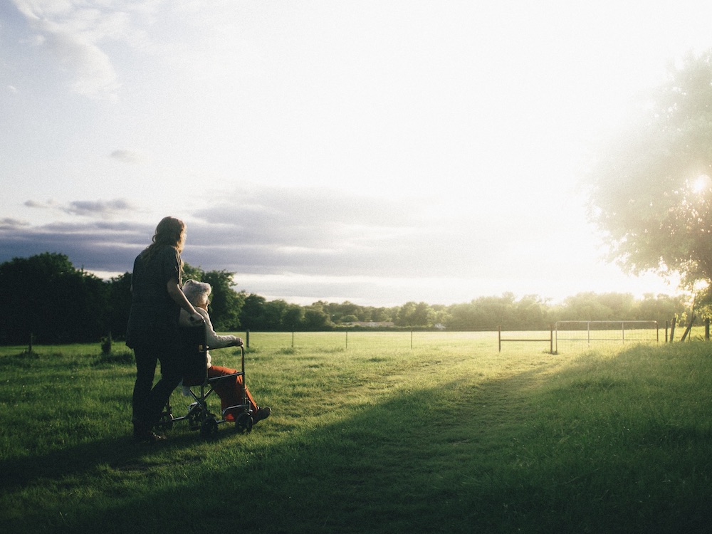 A senior is pushed in a wheelchair across a green grassy field towards the light of the sun.