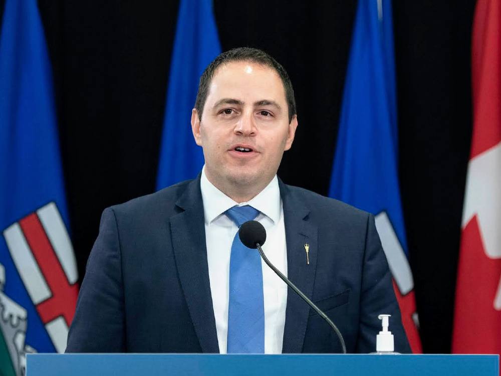 A man with short dark hair, blue suit and tie wearing a white shirt stands at a podium.