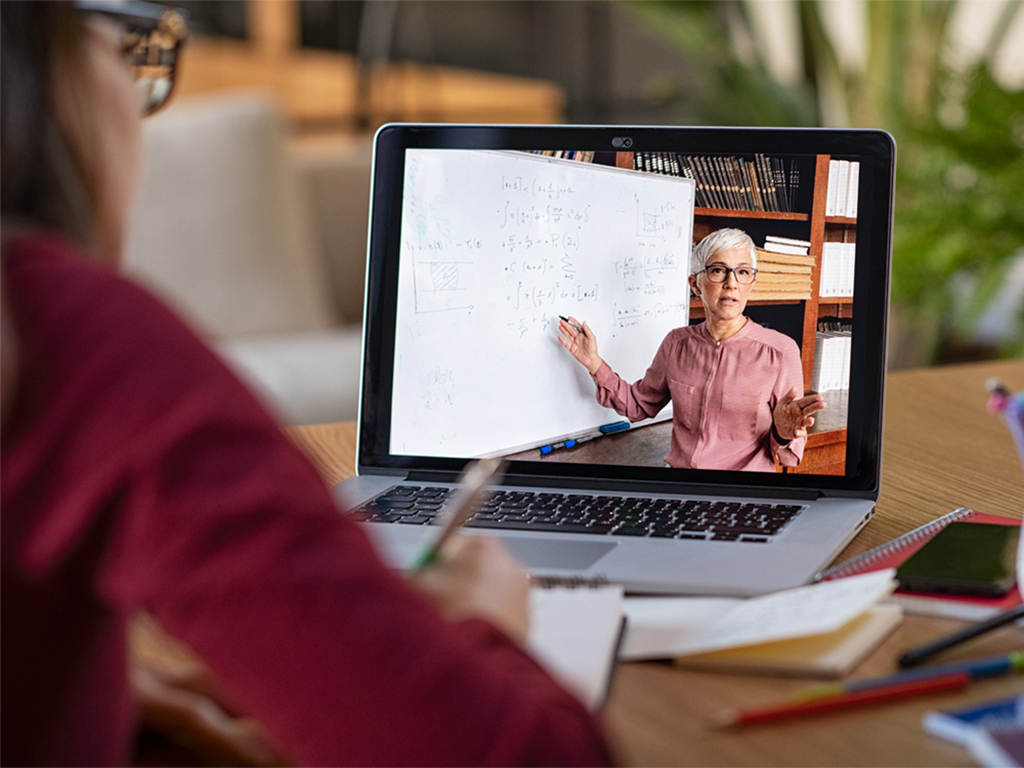 To the left, in profile, a student looks at a laptop. On the laptop, a professor with close-cropped grey hair teaches a math lecture.