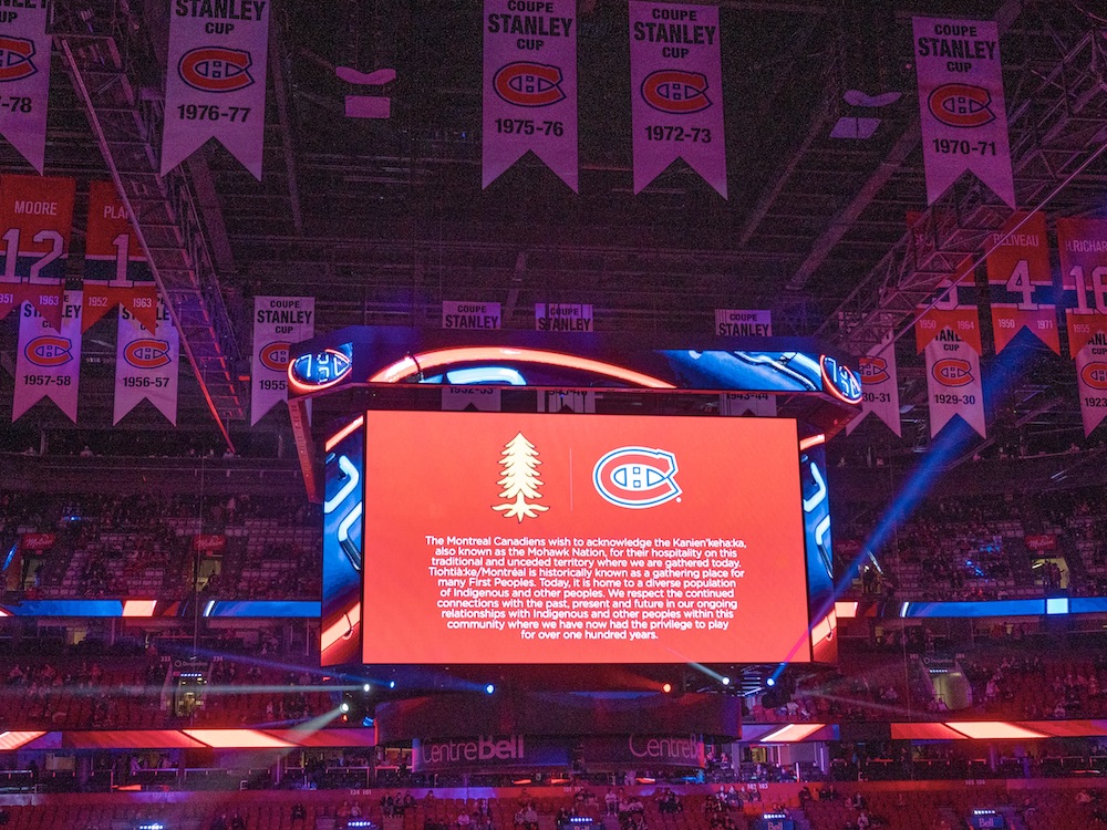 In a hockey arena, a land acknowledgement appears on the scoreboard above centre ice.