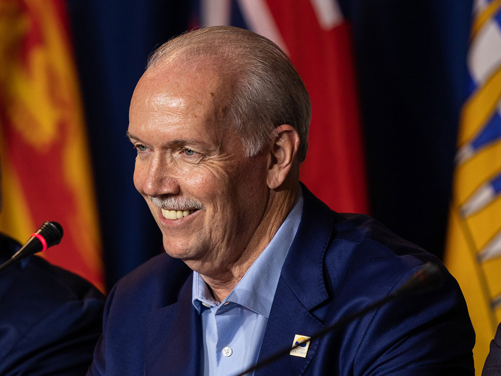 A smiling middle-aged white man with wrinkled eyes, greyish hair and a light blue shirt and darker blue jacket stands at a podium.