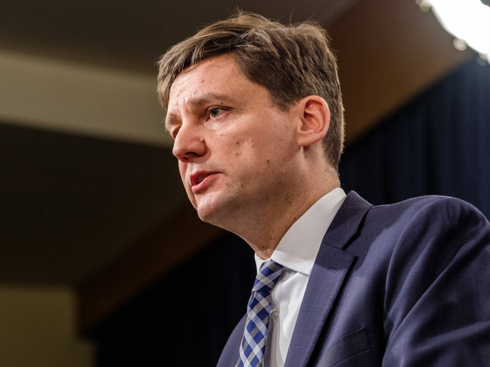 A light-skinned man with short scruffy hair stands at a podium wearing a dark blue suit, light shirt and blue patterned tie.