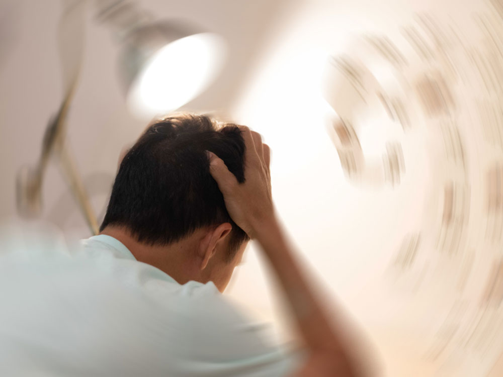 A dark-haired white man seen from behind clutches his head, a bright lamp and room out of focus around him.