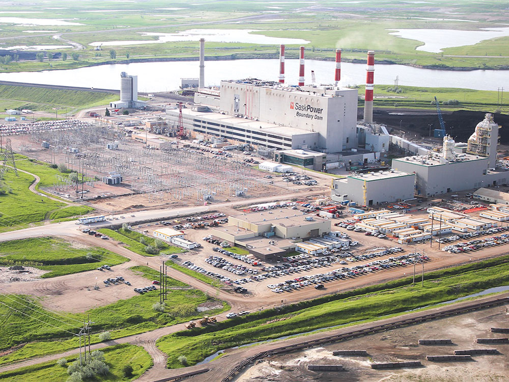 A large grey multi-storey windowless building is at the centre of an industrial complex, with four large rd and white smokestacks rising high into the air. In the foreground is electrical equipment and industrial trailers; in the background are cooling ponds.